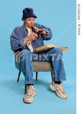 Interesting game. Cheerful man in jeans clothes sitting with pizza and playing retro game console over blue studio background. Concept of emotions, lifestyle, hobby 98599529