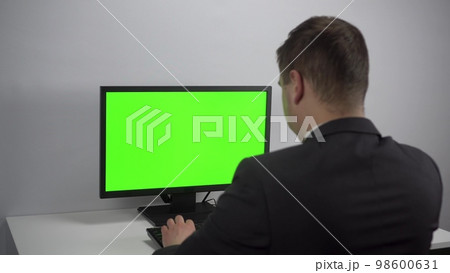 A young businessman is sitting in front of a computer with a green screen. A man in a suit works for a PC. Screen with chroma key. A young businessman is sitting in front of a computer with a green screen. A man in a suit works for a PC. Screen with chroma key. 98600631