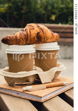 Fresh baked chocolate croissant on Two paper cups with lid for tea to go. Empty paper notebook. Freelancer's place of work. Study and work online. Remote business education. Making plans for next week 98603713