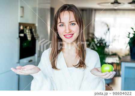 Portrait of young smiling woman holding green fresh apple in one hand, pill, vitamins in another, trying to decide which choice is the best one stand on kitchen. Diet modification, drugs alternative. 98604022
