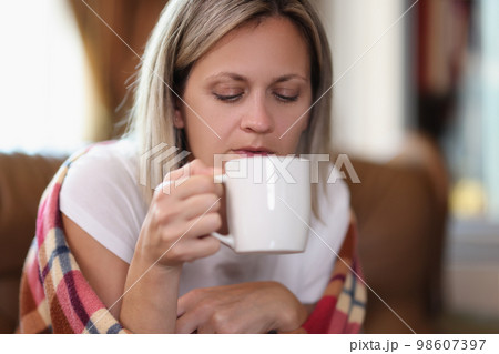 Freezing woman wrapped in warm checkered plaid blanket holds mug and drinks hot beverage at home. Freezing woman wrapped in warm checkered plaid blanket holds mug and drinks hot beverage at home. 98607397