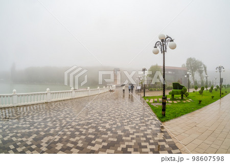 Beautiful wet and foggy embankment with people walking under umbrellas. Foggy rainy autumn landscape with modern park in Zheleznovodsk. 98607598