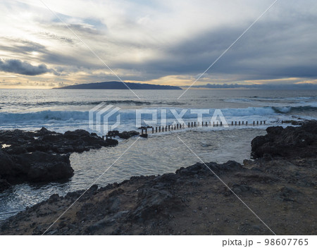View of the natural sea rock pool on the Tenerife coast near Alcala with vawes crushing against concrete wall and black volcanic rocks. Evening sunset sky. 98607765