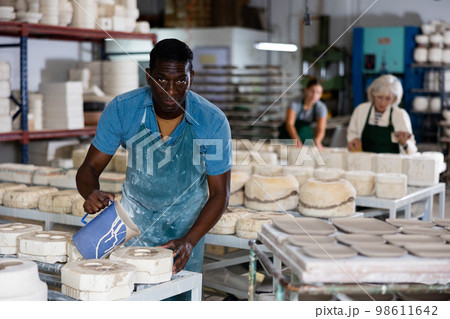 Slip casting - african american worker pours slip mass from jug into plaster mold in pottery factory 98611642