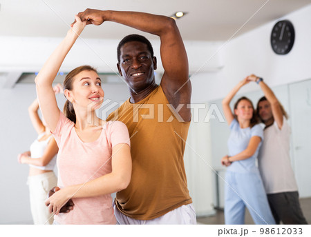 Positive smiling dancing couple enjoying active dancing during group training in dance hall Positive smiling dancing couple enjoying active dancing during group training in dance hall 98612033