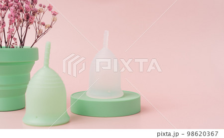 Menstrual cups on green podium next to red dry flowers in power case against pink background 98620367