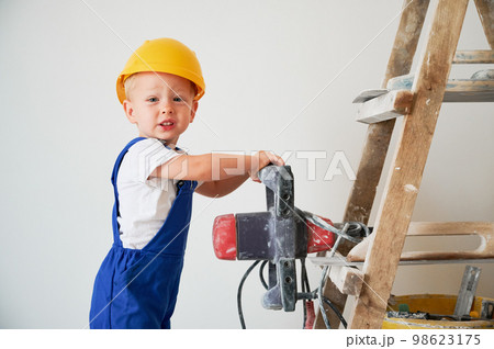 Adorable little boy construction worker holding electric power drill and looking at camera. Cute child wearing work overalls and safety helmet while standing on ladder against white wall. Adorable little boy construction worker holding electric power drill and looking at camera. Cute child wearing work overalls and safety helmet while standing on ladder against white wall. 98623175