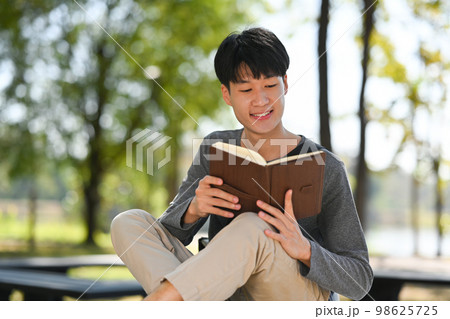 Happy young asian man reading interesting book while sitting bench in sunny summer park Happy young asian man reading interesting book while sitting bench in sunny summer park 98625725