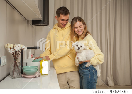 Portrait of happy couple in yellow hoodies, man washing the dishes, woman holding little white dog 98626365