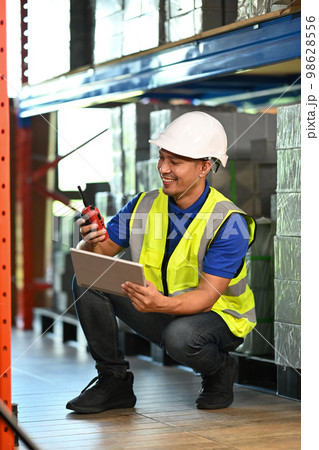 Warehouse worker using digital tablet, checking quantity of storage product in aisle between shelves full of packed boxes 98628556