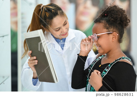 Happy child girl testing new glasses with optometrist and looking in the mirror at optical shop. Eyesight and vision concept 98628631