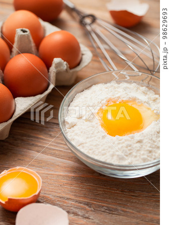Bowl plate with flour and egg yolk on wooden board with fresh raw eggs and metal whisk 98629693