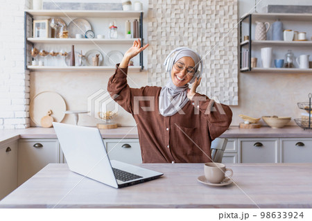 Young arab woman businesswoman, hijabi freelancer working from home online at home using laptop. She sits with headphones, listens to music, raised her hands, dances, rests, takes a break. 98633924