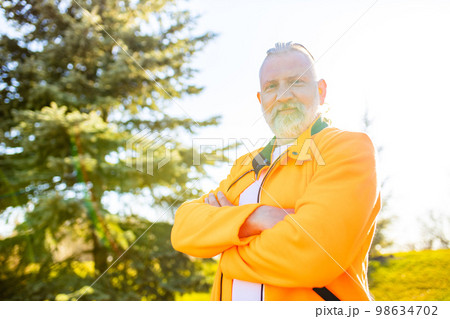 Senior man in orange sports wear doing stretching warming up outdoors sunny day in forest alone Senior man in orange sports wear doing stretching warming up outdoors sunny day in forest alone 98634702