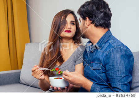 Young indian couple sitting in they living room and eating vegetable salad Young indian couple sitting in they living room and eating vegetable salad 98634769