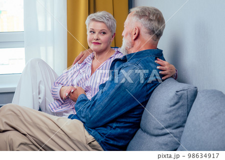 Mature good-looking well dressed couple sitting in sofa in day light from big window and yellow curtains in living room,touching hand 98634917