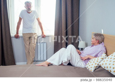 blonde shorh haircut woman quarreling with her elderly husband in day light bedroom.handsome man standing at the windowsill and looking at wife blonde shorh haircut woman quarreling with her elderly husband in day light bedroom.handsome man standing at the windowsill and looking at wife 98635040