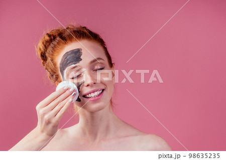 redhaired ginger teenager girl remove with cotton black clay mask on her pretty face on pink studio background 98635235