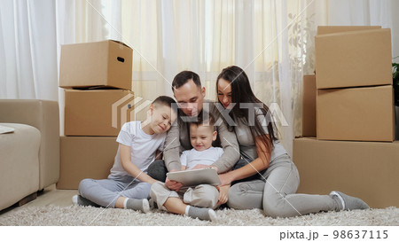 Excited family sits among boxes on floor and looks at tablet Excited family sits among boxes on floor and looks at tablet 98637115