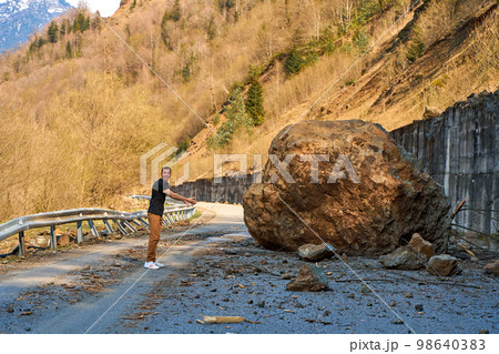 A huge rock fell from the mountains onto the road, destroying the asphalt and blocking half of the roadway 98640383