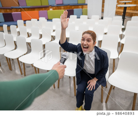 A red-haired Caucasian business woman sits in the front row in an empty conference room and answers into an outstretched microphone.  98640893