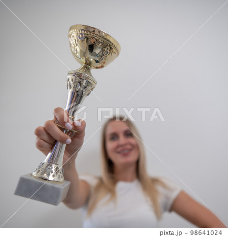 Blonde woman smiling and holding a golden trophy cup against a white wall. Blonde woman smiling and holding a golden trophy cup against a white wall. 98641024
