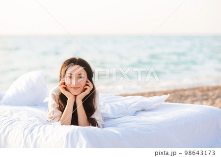 Smiling beautiful brunette woman lying in bed on white duvet with pillows over blue sea at background. Look at camera. Summer vacation season. 98643173