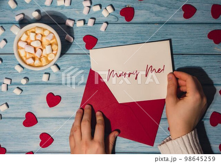 MARRY ME text on valentine card inscription positive quote phrase Female hands holding valentine card red envelope with white cup of coffee and marshmallows on wooden blue background. Romantic Small MARRY ME text on valentine card inscription positive quote phrase Female hands holding valentine card red envelope with white cup of coffee and marshmallows on wooden blue background. Romantic Small 98645259