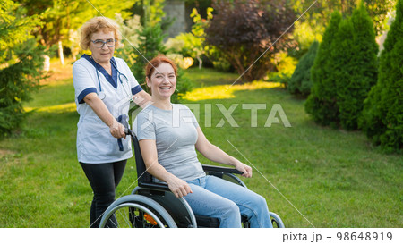 An elderly female nurse walks with a middle-aged woman in a wheelchair in the park.  98648919