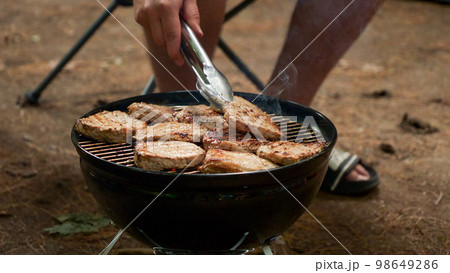 Unrecognized man is cooking BBQ stakes on charcoal grill in the forest. Camping food preparation. Fresh juicy stakes grilling on the barbeque. 98649286
