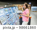 A beautiful smiling woman stands in a large grocery store near a grocery cart and chooses a holiday cake. A woman is holding a box of cake while standing near a refrigerator in a store. Shopping 98661860