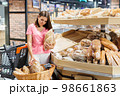 A beautiful smiling woman stands in a large grocery store near a grocery cart and chooses fresh fragrant bread. A woman stands near a basket of bread. Shopping concept 98661863