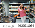 A beautiful woman stands in a large grocery store with a grocery cart near shelves with alcoholic beverages. A woman stands near the shelves with bottles. Shopping concept 98661864
