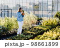 A young woman stands outside a plant shop and chooses a pot with a small tree. A woman chooses plants for landscaping the yard. Home gardening concept 98661899