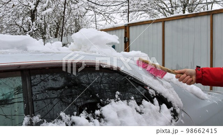 the hand of a person in a red jacket cleans snow from the roof and windows of a gray car with a brush in a winter yard under a cloudy sky, cleaning snow drifts from the car body 98662415