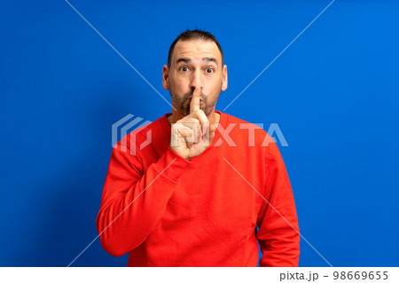 Serious handsome bearded hispanic man puts his index finger to his lips, tries to keep conspiracy, looks aside and says: Shh, be quiet please. Isolated shot of attractive man shows silence sign. 98669655