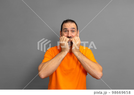 Latino man with a beard in an orange t-shirt biting his nails out of fear, he is experiencing a distressing situation. Isolated on gray background. 98669658