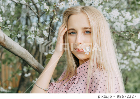 Blonde girl on a spring walk in the garden with cherry blossoms. Female portrait, close-up. A girl in a pink polka dot dress. 98672227
