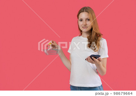 Beautiful caucasian young woman holding small shopping cart and mobile phone Beautiful caucasian young woman holding small shopping cart and mobile phone 98679664
