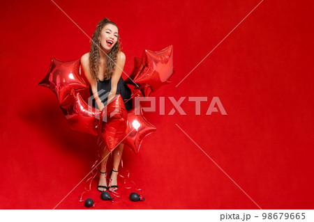 Beautiful young joyful girl is holding bunch red star balloons on red 98679665
