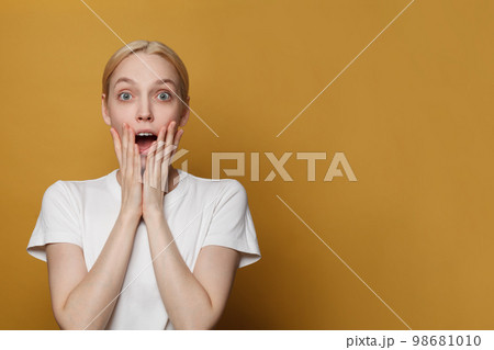 Surprised screaming blonde woman in white t-shirt looking at the camera over yellow studio wall 98681010