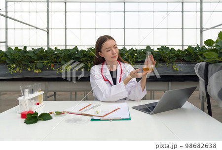 Caucasian female botanical scientist in white gown observes yellow chemical in Erlenmeyer flask. The table is full of research papers, laptop computer, chemicals in test tubes and plant samples. Caucasian female botanical scientist in white gown observes yellow chemical in Erlenmeyer flask. The table is full of research papers, laptop computer, chemicals in test tubes and plant samples. 98682638