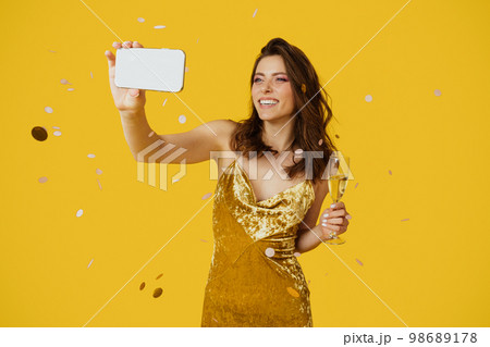 Happy woman in stylish dress taking selfie with glass of champagne, yellow studio background with falling confetti 98689178
