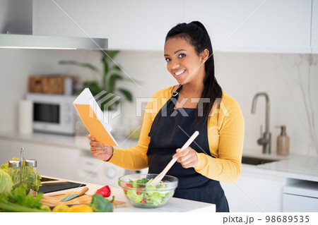 New recipe and homemade food. Happy millennial african american lady in apron make salad, read notebook New recipe and homemade food. Happy millennial african american lady in apron make salad, read notebook 98689535