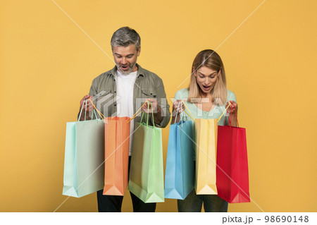 Shopaholics. Overjoyed middle aged caucasian couple holding shopping bags and looking inside, yellow background Shopaholics. Overjoyed middle aged caucasian couple holding shopping bags and looking inside, yellow background 98690148