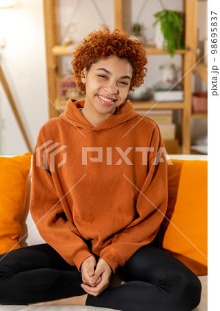 Beautiful african american girl with afro hairstyle smiling sitting on sofa at home indoor. Young african woman with curly hair laughing. Freedom happiness carefree happy people concept Beautiful african american girl with afro hairstyle smiling sitting on sofa at home indoor. Young african woman with curly hair laughing. Freedom happiness carefree happy people concept 98695837