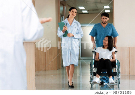 Doctor and nurse transport female patient in wheelchair at sterile corridor. 98701109