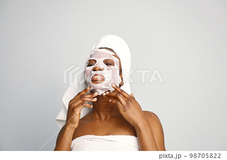 Black young woman standing isolated on white background with sheet mask on her face. Girl has her hair wrapped in a towel. Black woman enjoy after body treatment. 98705822