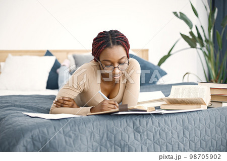 Young black woman lying down on bed reading a book at home. Girl wearing eyeglasses and beige sweater. Girl preparing her homework. Young black woman lying down on bed reading a book at home. Girl wearing eyeglasses and beige sweater. Girl preparing her homework. 98705902