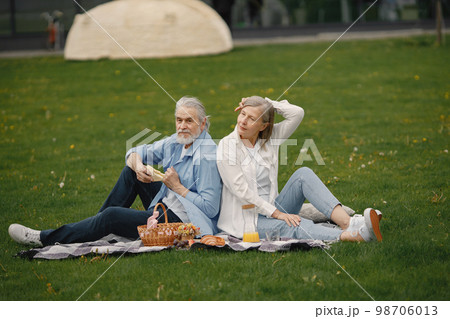 Beautiful caucasian elderly couple sitting on a grass in the park in summer. Man and woman sitting on blanket at the park drinking juice and sharing few precious memories. Woman wearing white shirt 98706013